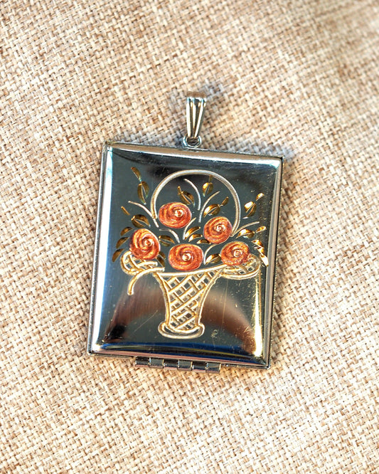 Silver-tone hinged locket pendant with enamel rose basket motif, closed, front view on neutral linen