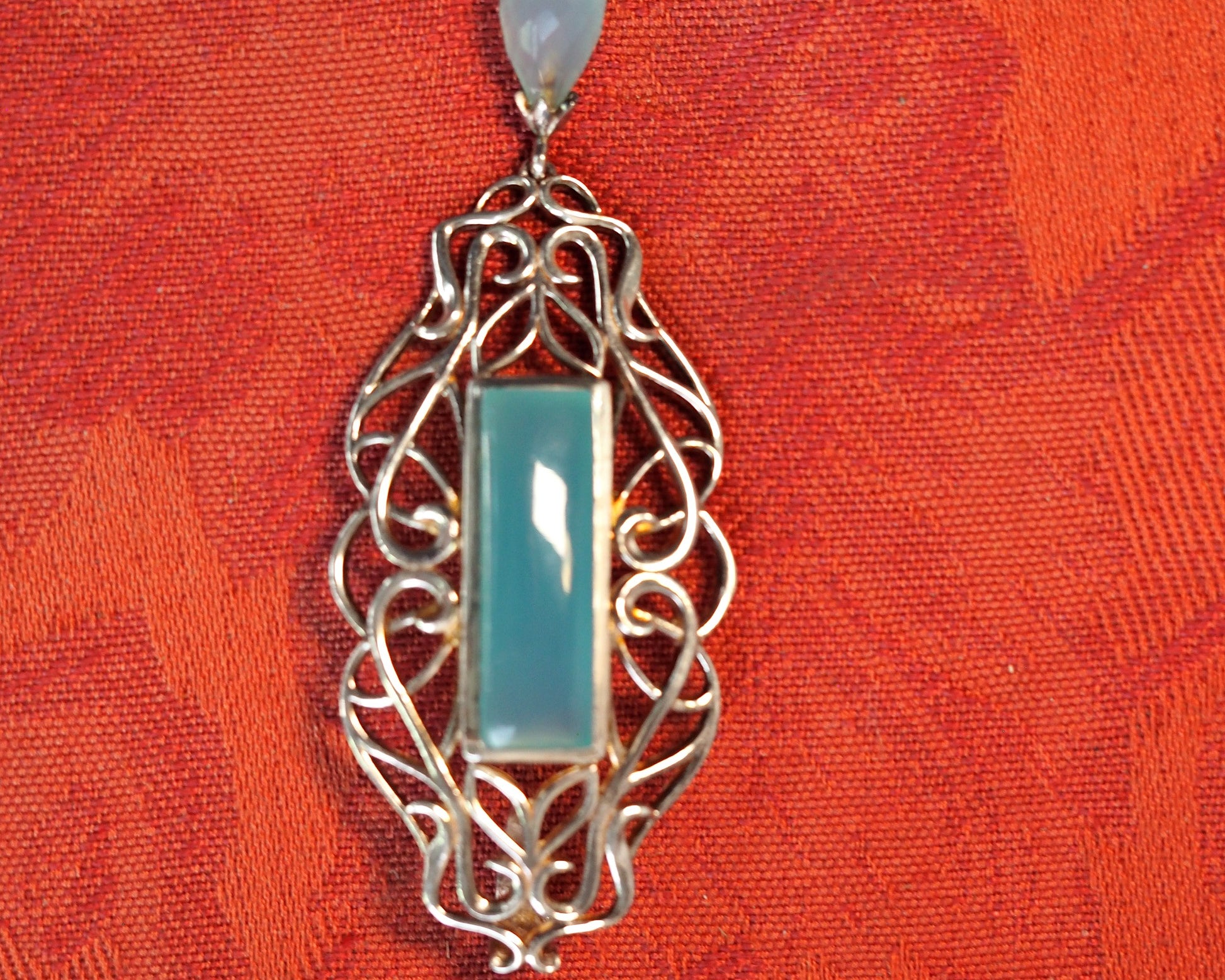 Derewala sterling silver and chalcedony pendant, front view flat on red fabric showing scrollwork frame