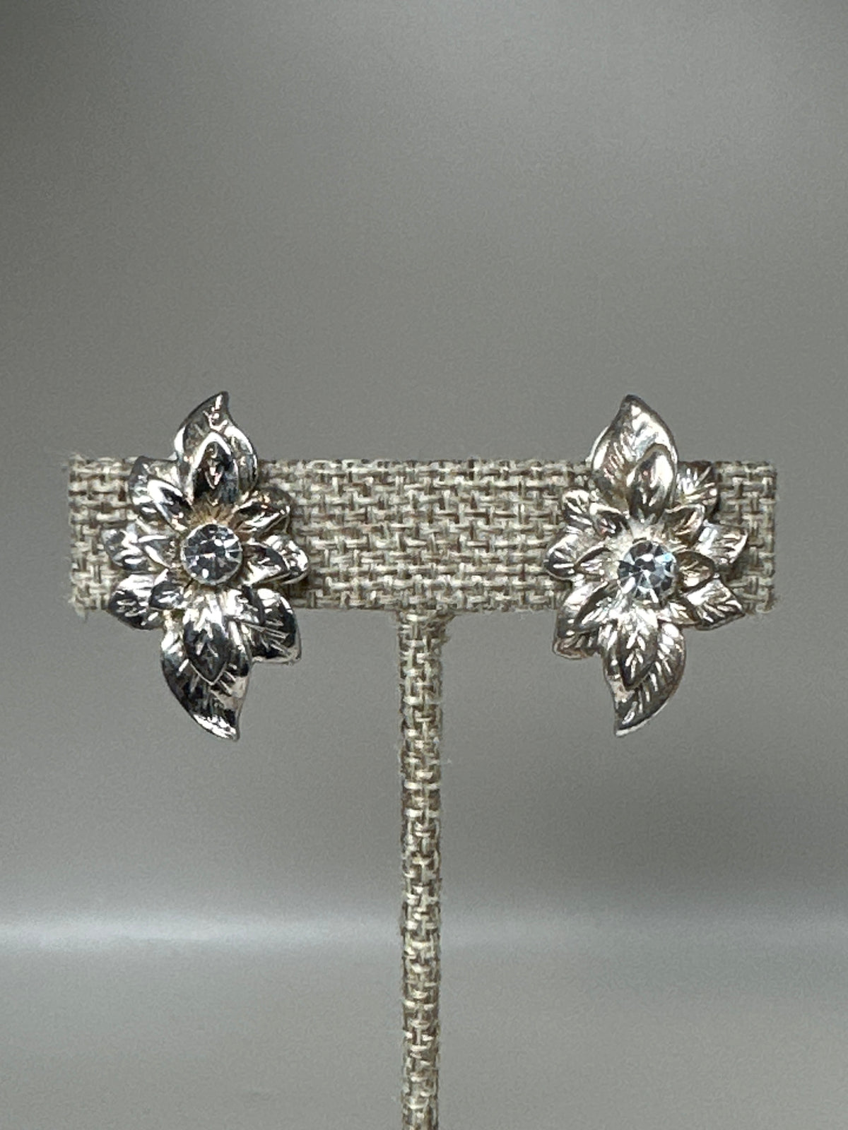 Silver-tone foliate spray clip earrings with rhinestone centers displayed on earring stand, front view