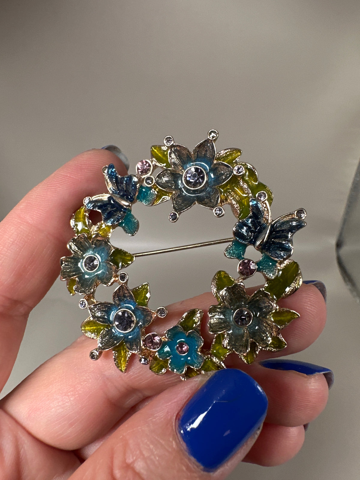 Napier enamel floral wreath brooch held in hand, showing six multi-color flower clusters with butterflies and rhinestone centers, front view