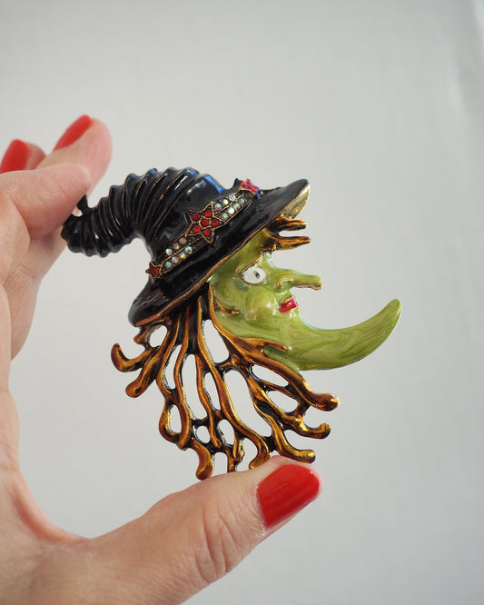 Kirks Folly witch brooch held in hand for scale, green enamel face, black hat and gold-tone fringe