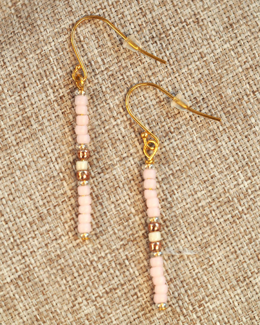 Pink Japanese glass bead drop earrings with gold-tone spacers on fishhook ear wires, shown flat on woven textile background