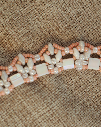 Detail view of beaded collar showing white cube beads with iridescent finish, gray ovals, and peach seed beads on burlap texture