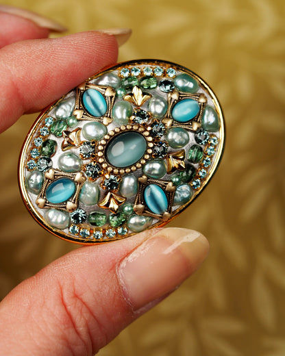 Michal Golan teal catseye brooch held between fingertips for scale, showing 1.75-inch oval size