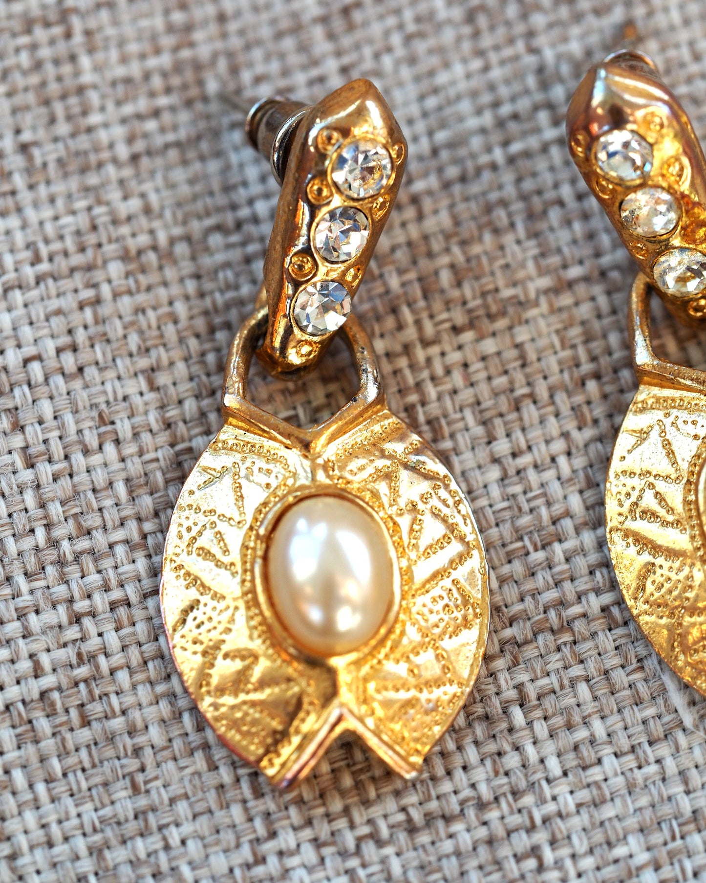 Close-up angle view of gold-tone leaf earrings showing stippled texture, faux pearl cabochon, and three rhinestones on curved post