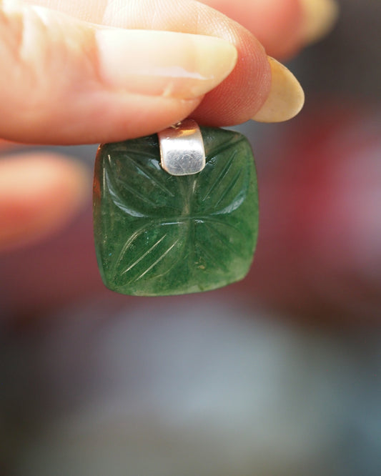 carved green aventurine pendant held against a fingertip, showing the floral relief pattern and silver bail, front view