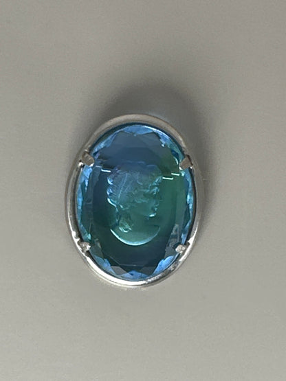 Blue glass cameo brooch, front view on flat surface, showing engraved geometric border on silver-tone metal frame