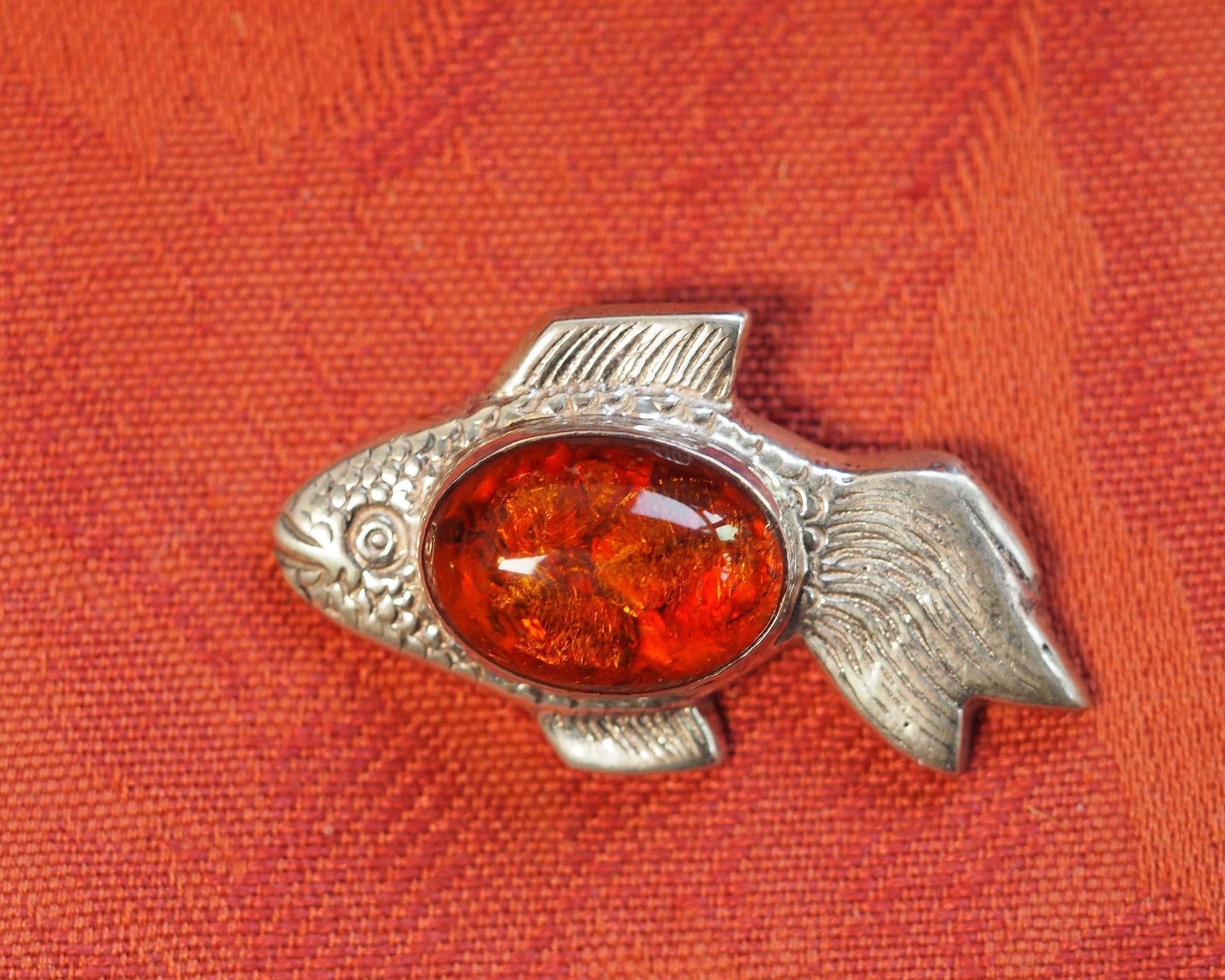 Fish pendant front view on orange fabric, showing textured scales, fins, and amber stone