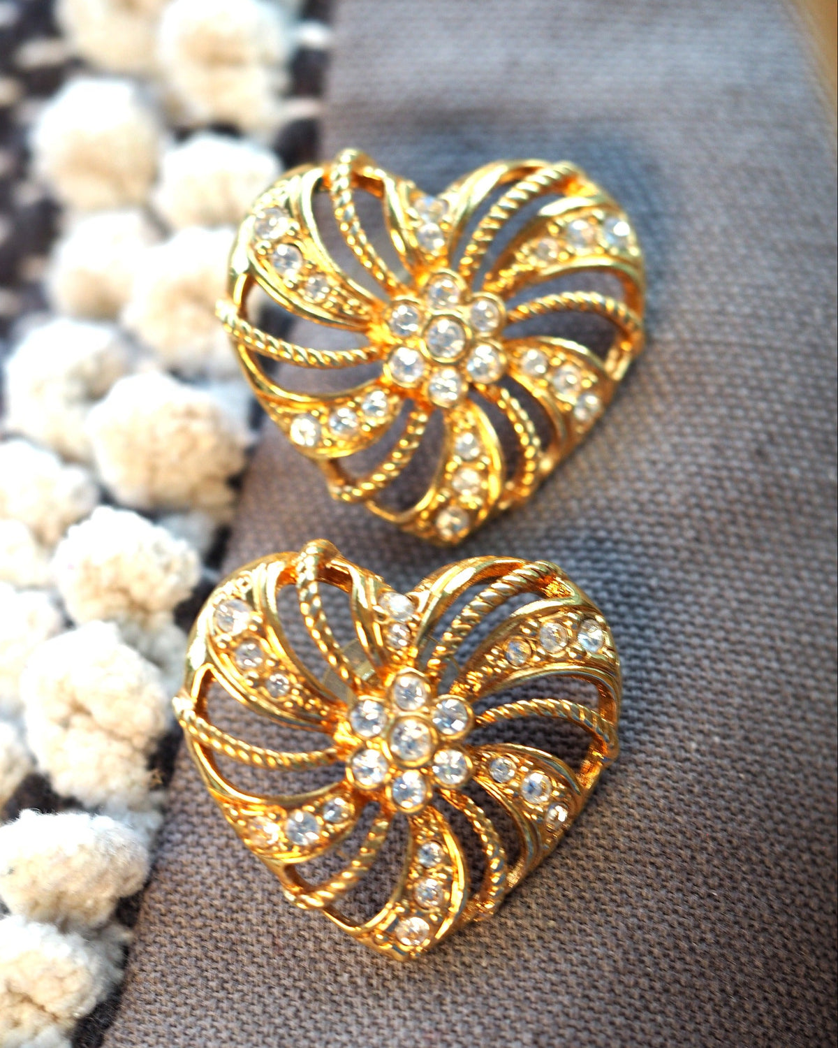 1980s gold-tone heart-shaped earrings with rhinestone-studded pinwheel petals, front view on gray fabric