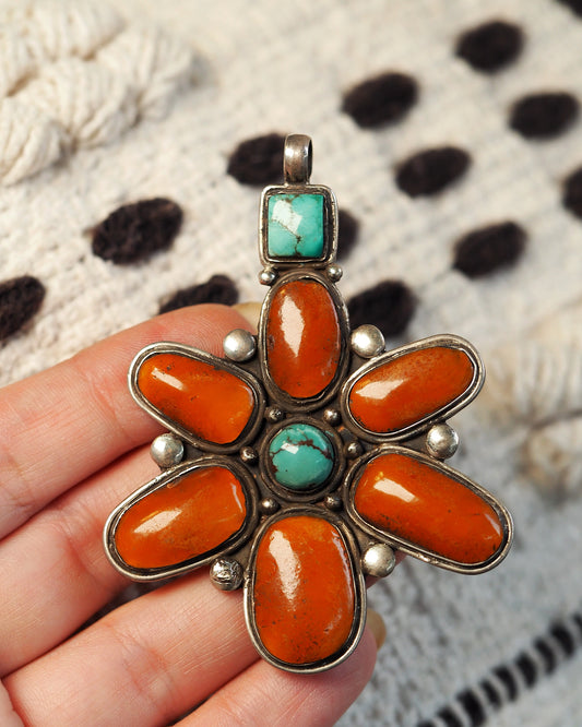 Sterling silver spiny oyster and turquoise flower pendant held on fingertips, showing six burnt-orange petals radiating from a central turquoise cabochon, front view