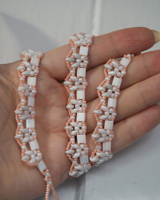 Close-up of necklace held in hand showing three bead shapes: peach seed beads, white iridescent cube beads, gray oval beads in dimensional rows