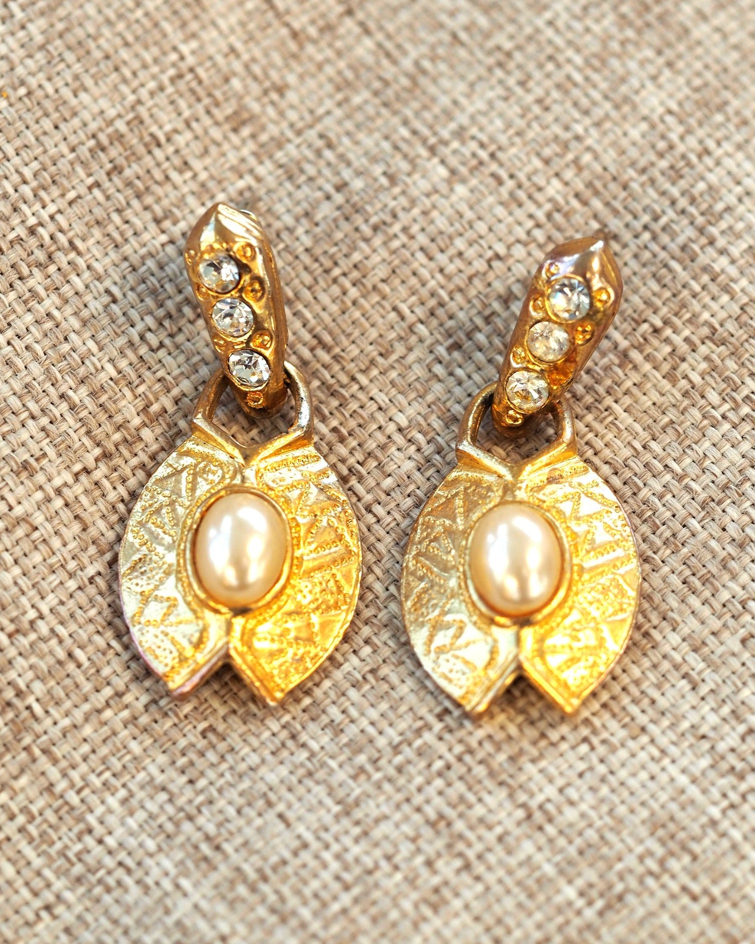 Gold-tone faux pearl leaf drop earrings with rhinestone accents on a neutral linen background, front view