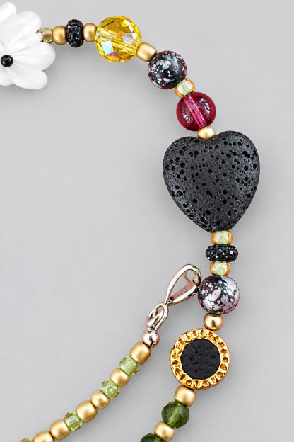 Close-up of black lava stone heart-shaped bead flanked by green seed beads, faceted glass beads, and gold-tone disc with black center