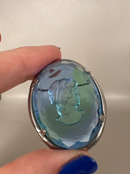 1960s blue glass cameo brooch held in fingertips, showing classical female profile carved in relief with green and purple color shifts