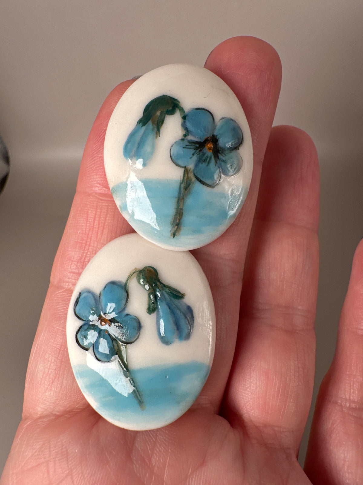 Close-up of ceramic earrings in hand showing scale and detail of hand-painted blue flowers with green foliage