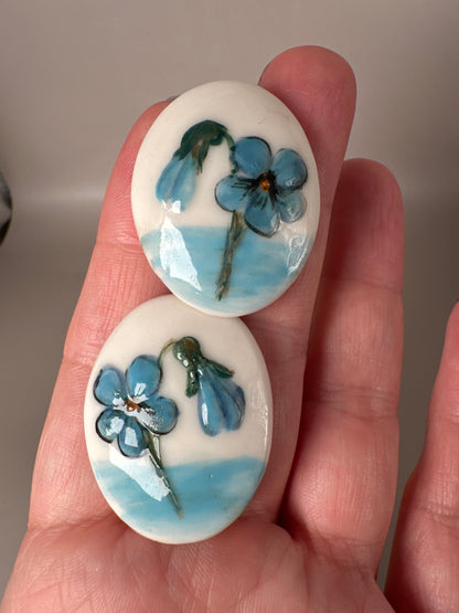 Close-up of ceramic earrings in hand showing scale and detail of hand-painted blue flowers with green foliage