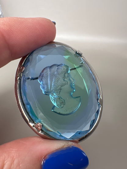 Blue glass cameo brooch held in fingertips at an angle, showing depth of the faceted glass and relief carving of the classical profile