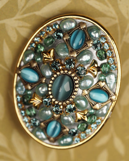 Close-up of Michal Golan brooch showing central teal catseye halo, fan flourishes, and faceted crystal border