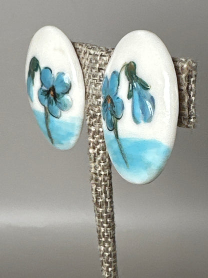 Pair of ceramic earrings showing hand-painted blue flowers and green leaves, side angle view
