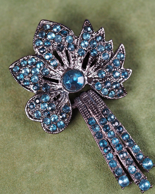 gunmetal-tone floral brooch with teal rhinestones in layered petals and articulated drop, front view on green surface