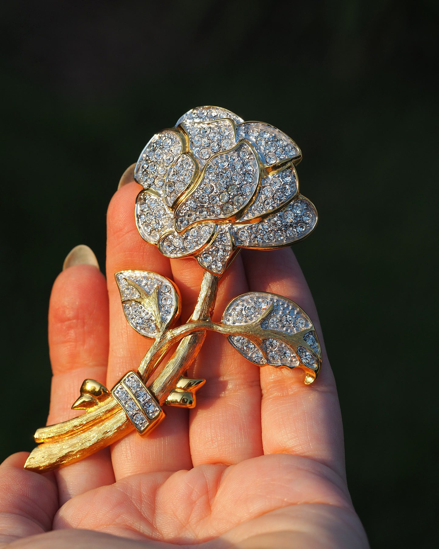 Nolan Miller gold-tone rhinestone rose brooch held in hand, front view showing layered petals and articulated leaves on brushed stem