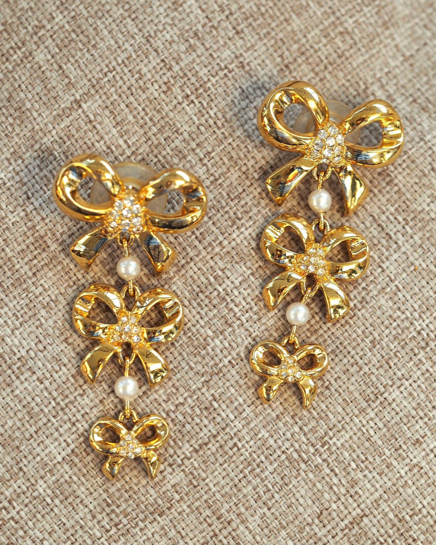 Pair of gold-tone bow drop earrings with rhinestone centers and pearl links, shown flat on linen, front view